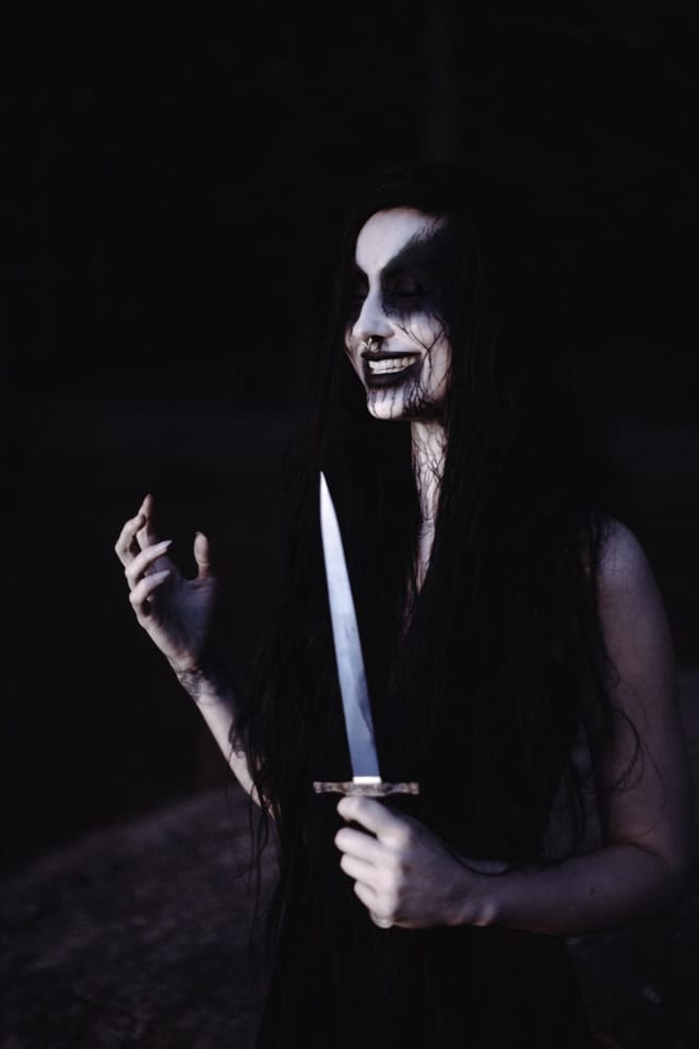 En leende black metal inspirerad gestalt med en ljus dolk mot en mörk skogsbakgrund