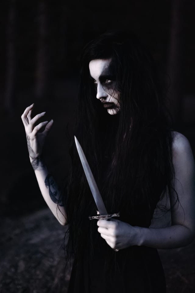 En black metal inspirerad gestalt som höjer ena handen intill en dolk i dämpat skogsljus