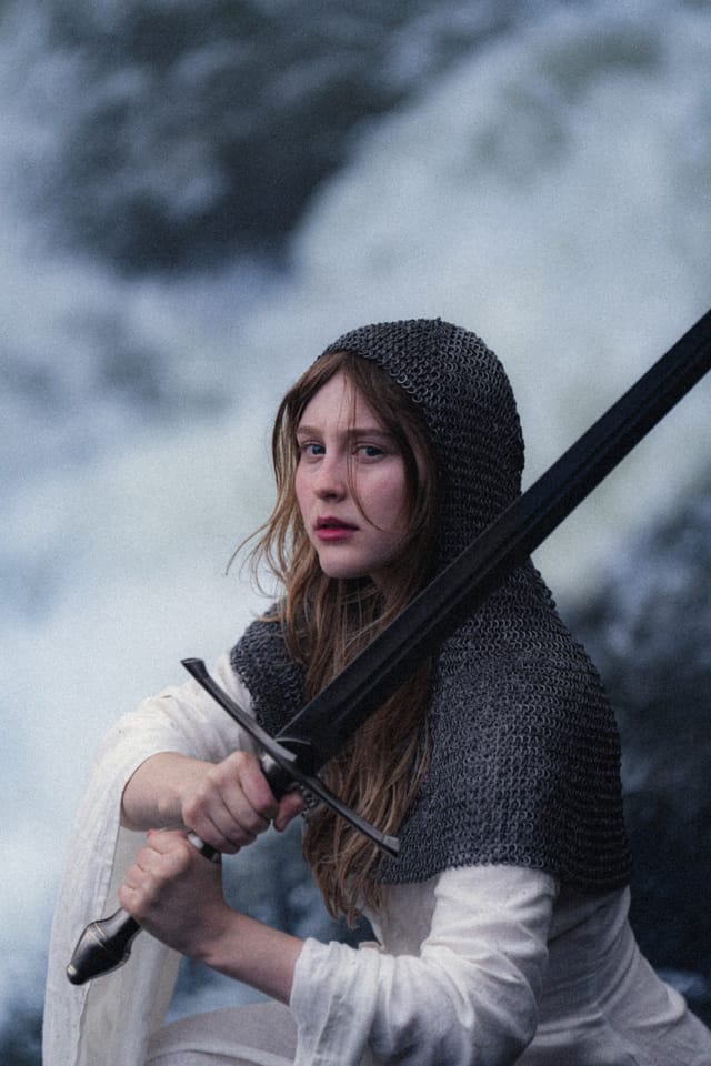 A chainmail-clad woman holding a sword diagonally before a blurred waterfall