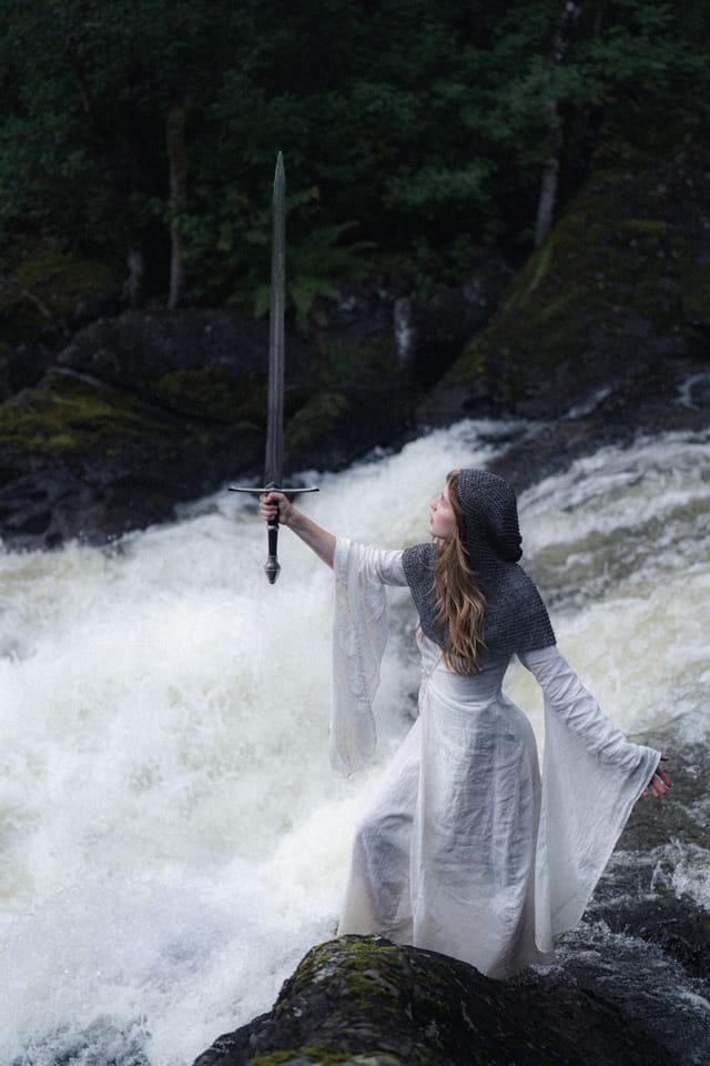 A woman in white raising a sword beside a rushing Norwegian waterfall