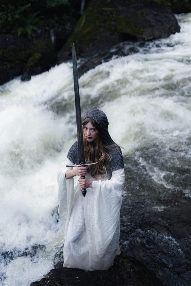 A sword-bearer in white standing on dark rocks amid foaming water