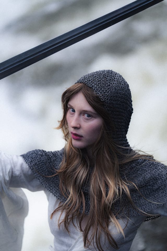 A close portrait beneath chainmail with a sword crossing the bright waterfall behind