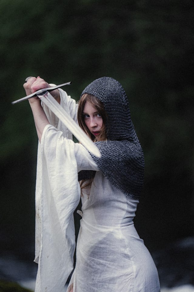 A woman in white lifting her sword against a deep green forest