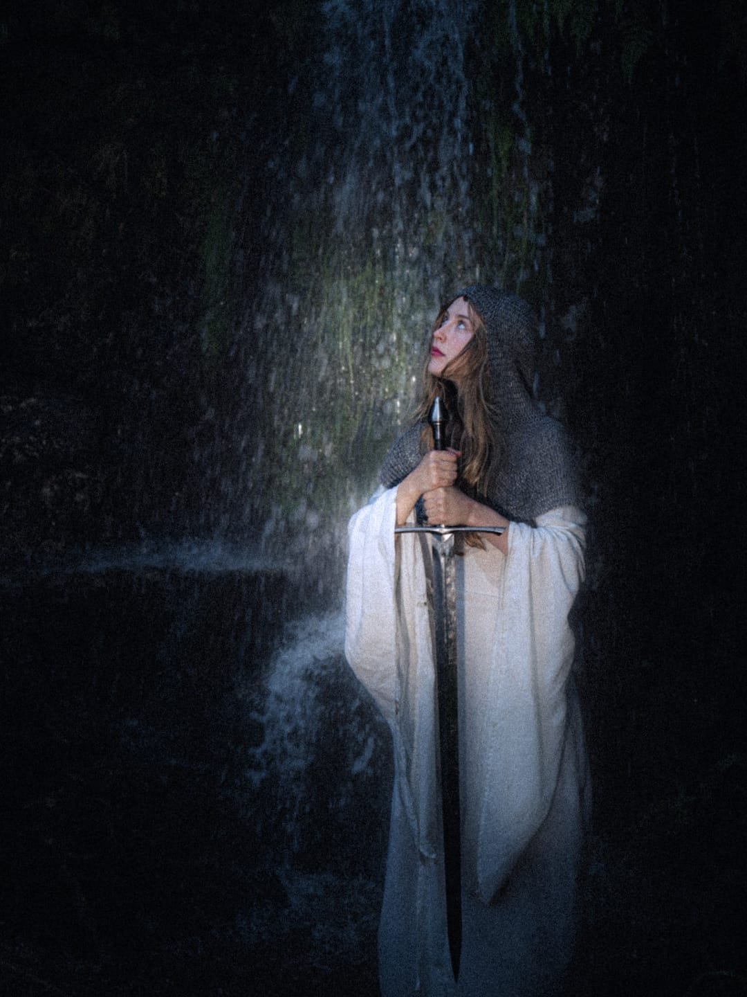 A white-robed woman resting a sword before a shadowed waterfall