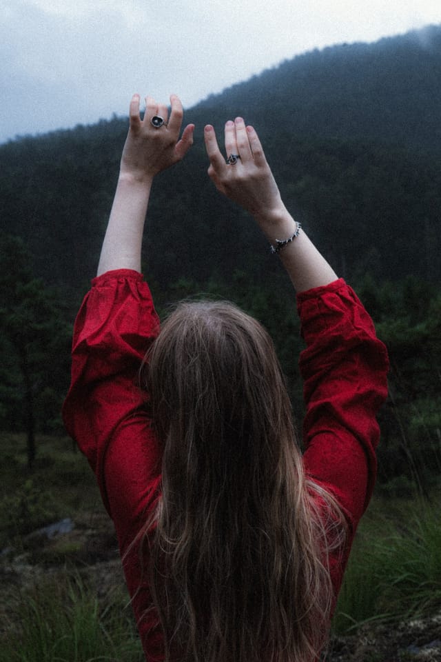 A woman in a red dress seen from behind with raised hands beneath misty hills