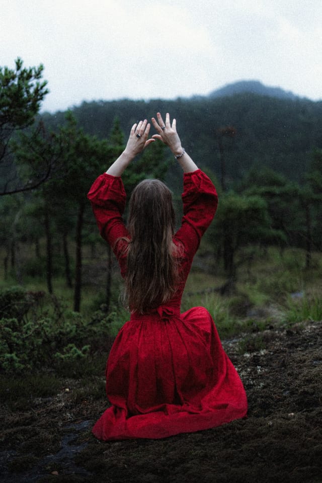A red-clad figure kneeling with her hands lifted toward the mountain mist