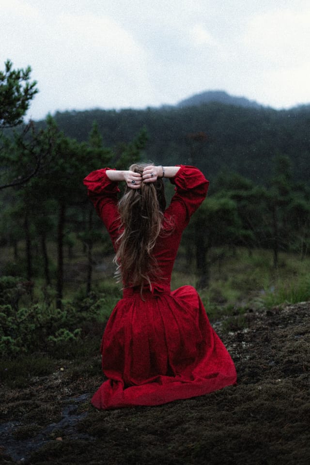 A red-clad woman resting both hands behind her head before dark hills