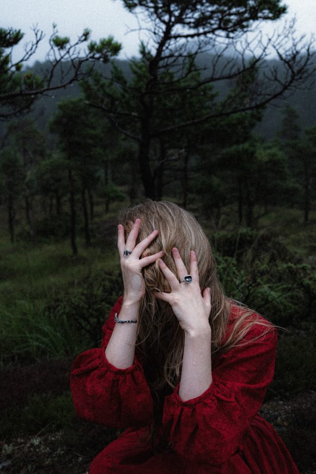 A woman in red covering her face with ringed hands in a dark woodland