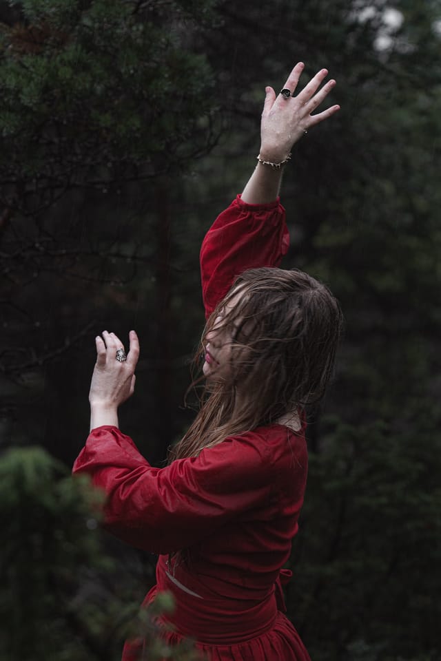 A woman in red turning in profile with her hands raised among dark trees