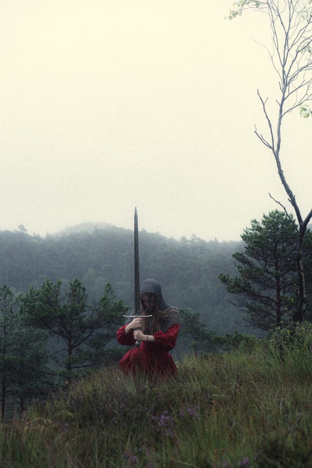 A woman in red standing with an upright sword before fog-covered hills