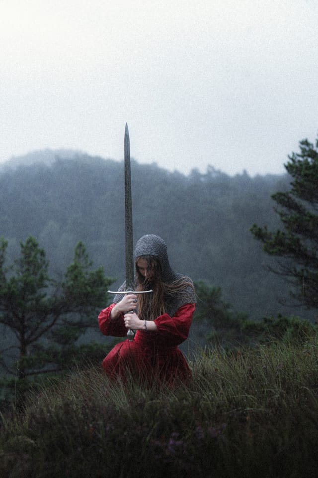 A full-length sword-bearer in red looking upward beneath a pale sky