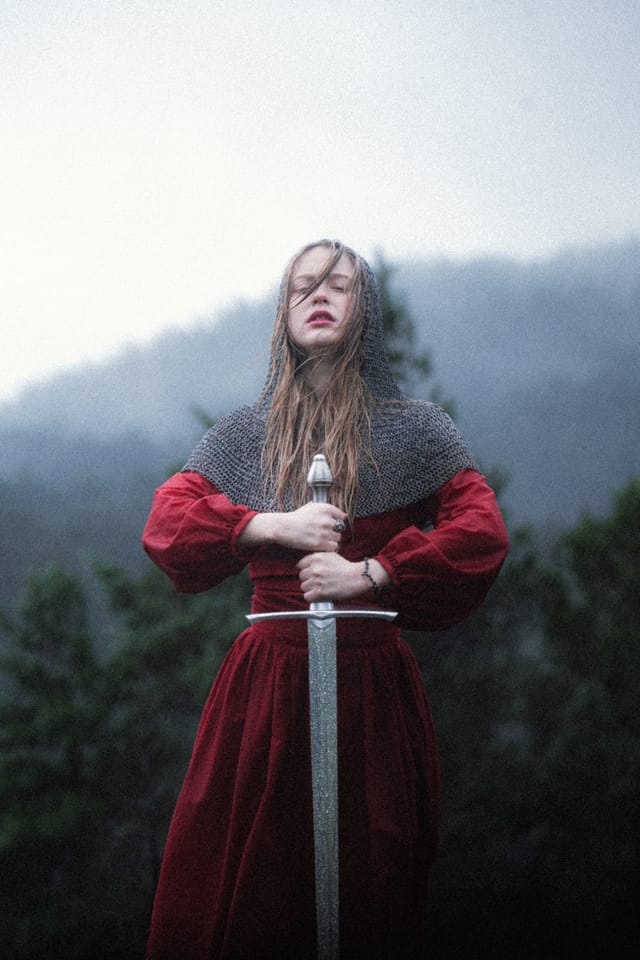 A chainmail-clad sword-bearer lifting her blade into the mountain mist