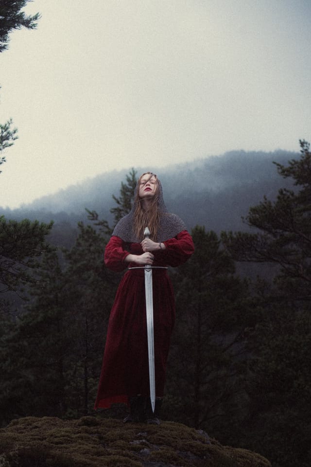 A woman in red holding a tall sword before a softly fogged hillside