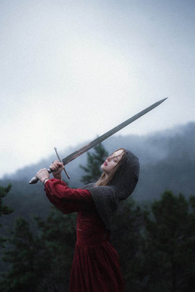 A woman in chainmail and red holding a sword across a pale mountain horizon