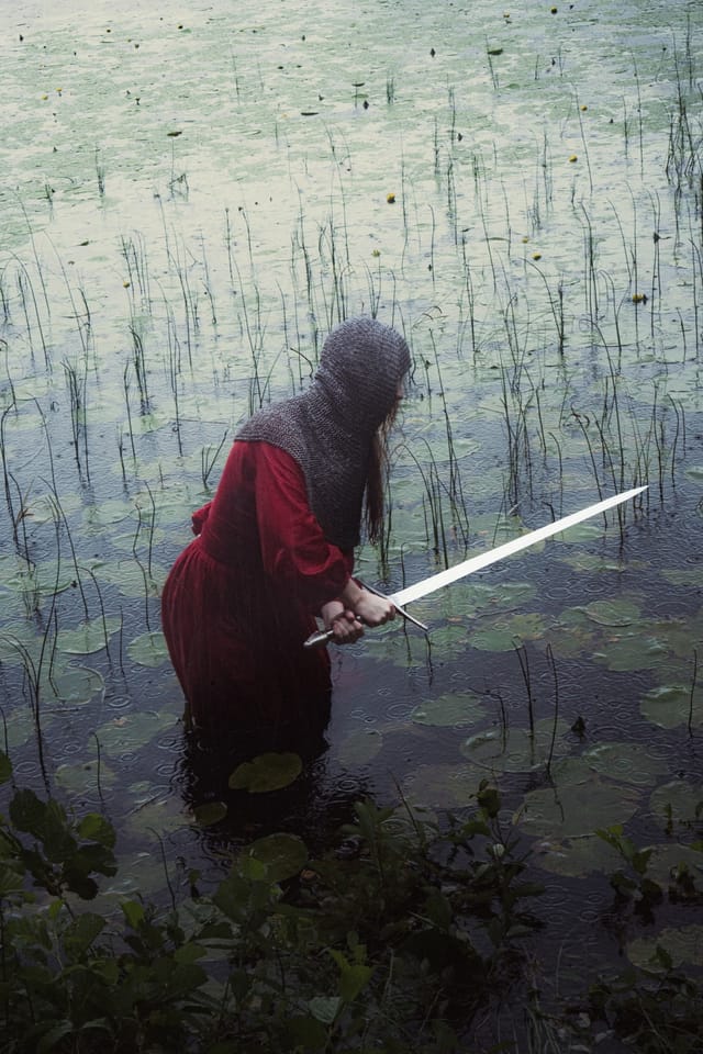 A red-clad sword-bearer wading through dark water and pale reeds