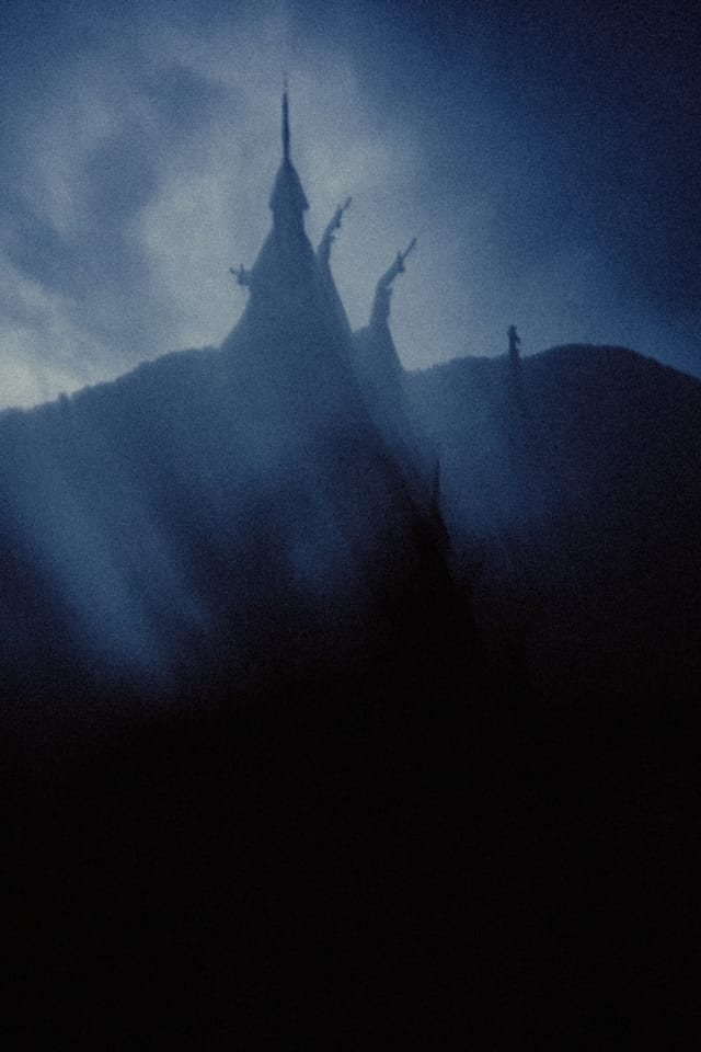 A stave church spire and carved roof peaks silhouetted above a dark ridge in storm-blue haze