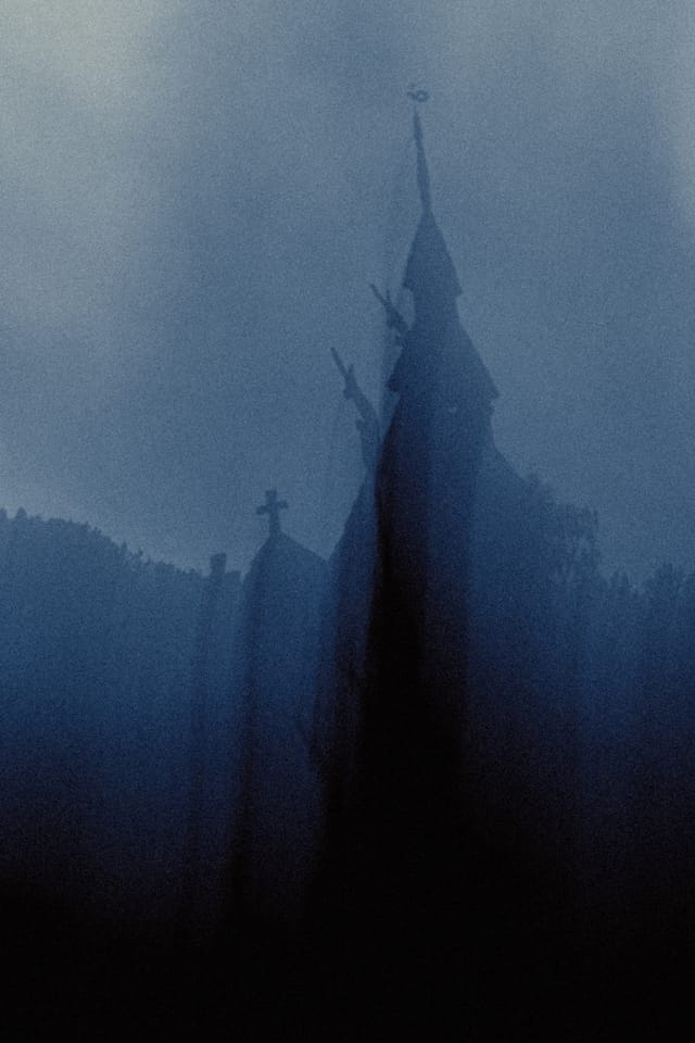 A narrow stave church tower emerging between dark rooflines in cold mountain fog