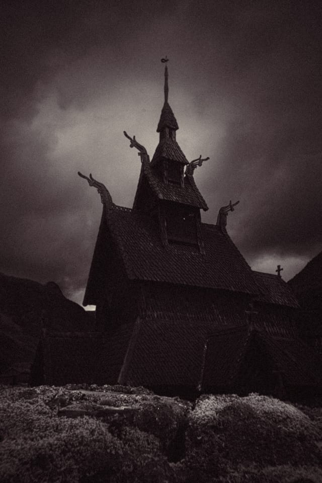 A dark stave church and carved gables standing above rocks under a dramatic sky
