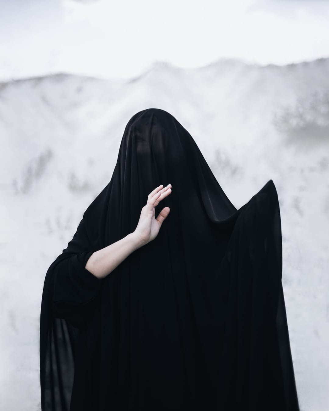 Cheri concealed beneath a black veil before a pale mountain landscape