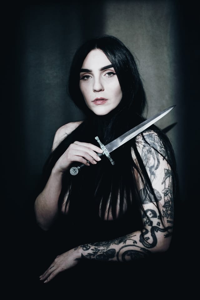 Cheri holding a dagger across her chest in a dark, softly lit portrait